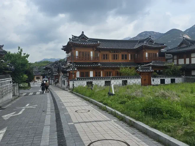 A grey paved road on the left with a traditional two-storey hanok on the right with a patch of grass in front of it