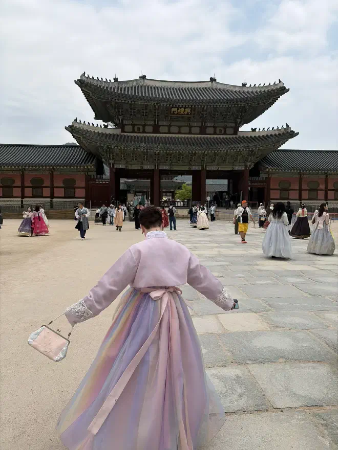 A person in a pink Hanbok with a pink/purple dress walking towards a traditional Korean palace entrance