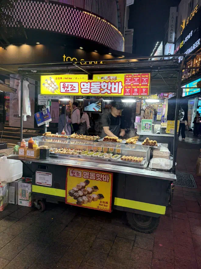 A man stands at a stall selling corn dogs covered with various toppings