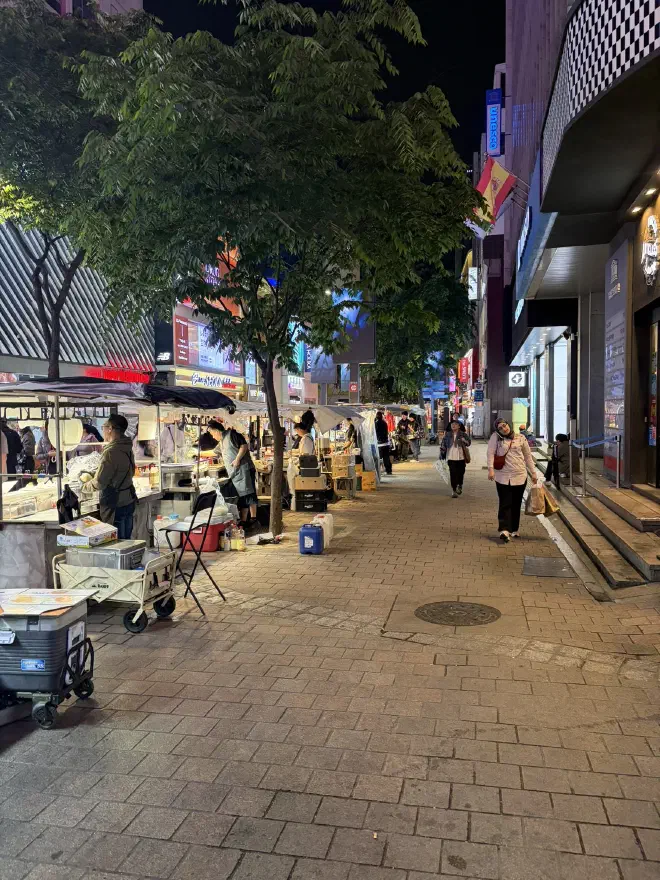 Food stalls line the side of a street lined with occasional trees at night