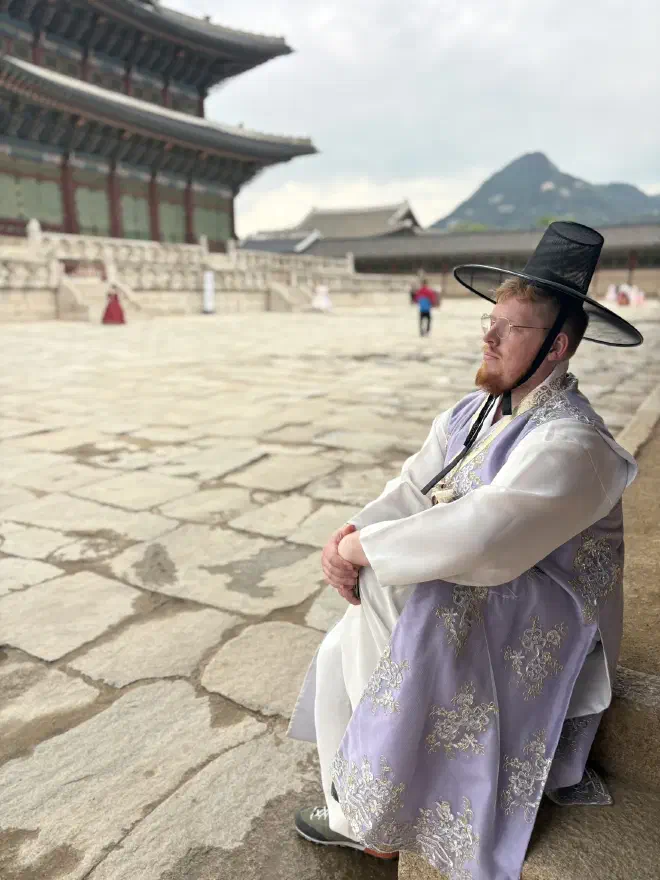 A man in purple jeogori, white trousers and a black wide-brimmed hat, sits in a stone courtyard with a traditional Korean building in the background
