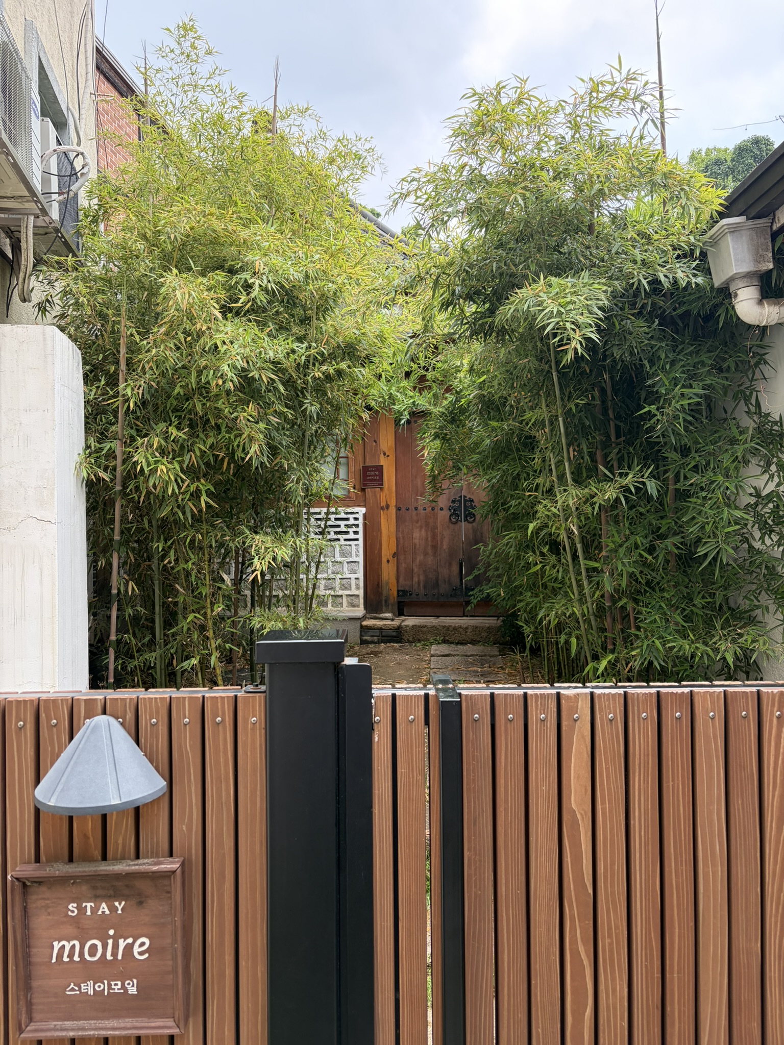 The Entrance to Stay moire A wooden gate and a sign saying ‘Stay miore’ and behind it a short path with bamboo either side leads to a wooden door