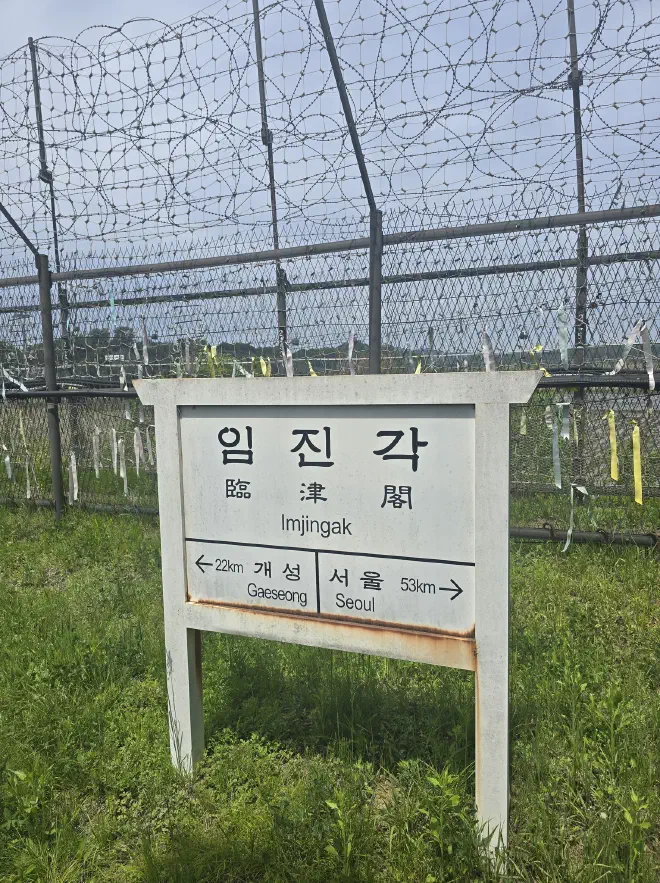 A white wooden sign in front of a barbed wire fence with an arrow pointing left (22km to Gaeseong) and an arrow pointng right (53km to Seoul).