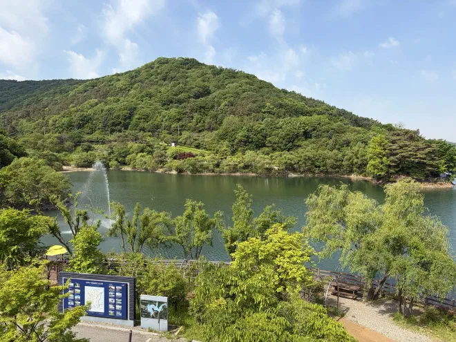 A view out over Majang Lake A tree covered hill behind a lake with a path running in front of it with an informational sign