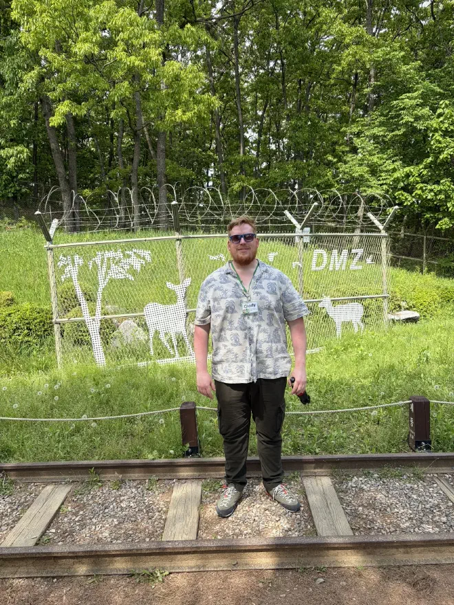 The photo opportunity fence at the DMZ A man in a flowery shirt standing on train tracks in front of a decorative barbed wire fence inscribed with the text 'DMZ'