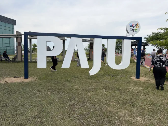A large sign with white letters saying 'Paju' on a dry lawn with people walking around it