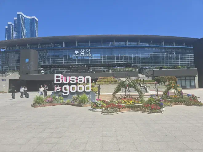 A long glass building with the text 'Busan Station' sits behind a small garden of flowers with a two meter tall 'Busan is good' sign