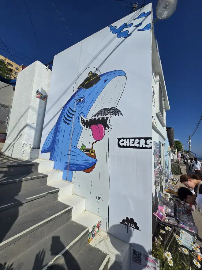A shark with a sailor had, a coconut with a straw, and a mustache, painted on the side of a shop while people browse products