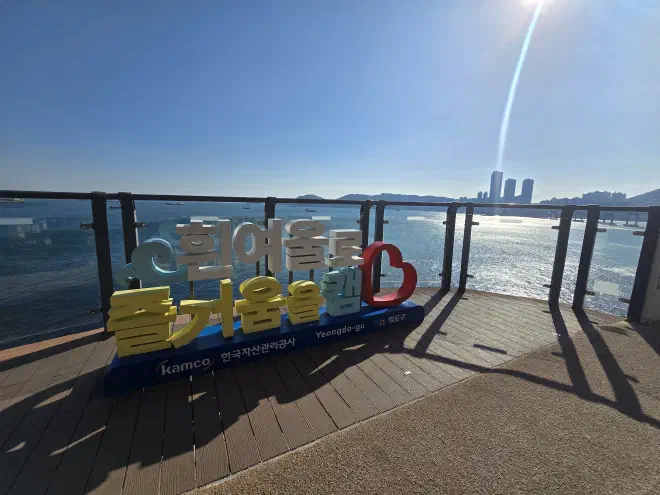 A wooden viewing platform with a glass wall looks out over the ocean with Busan in the background