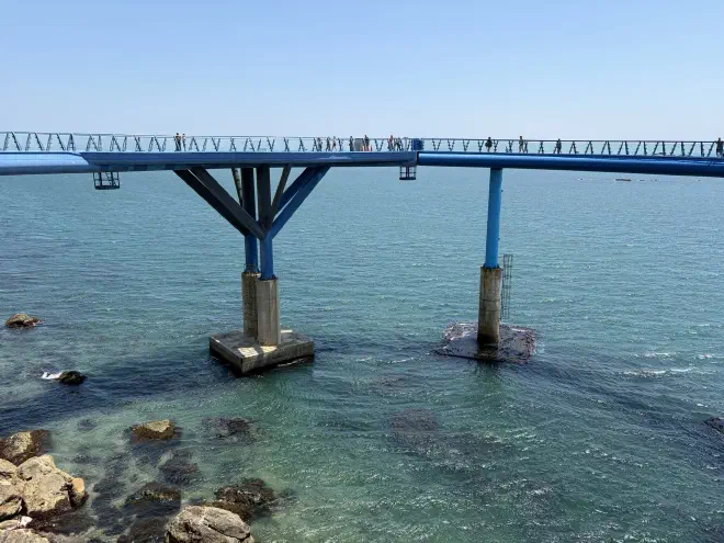 A blue metal walkway on posts sits above the ocean with people walking along it