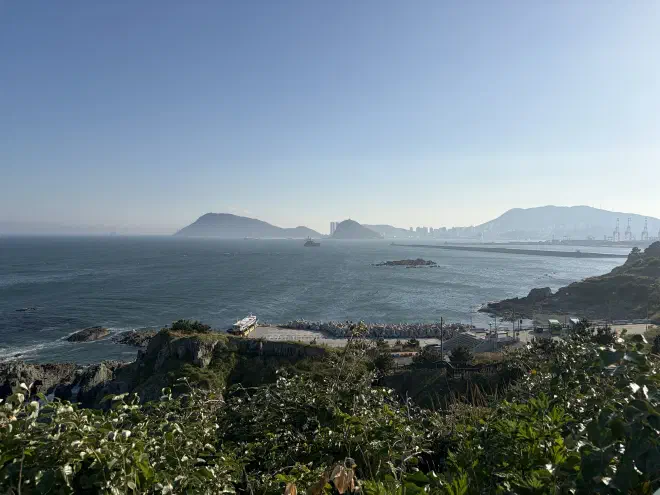 View west from the Oryukdo Skywalk An ocean with a city and mountains in the distance and a concrete dock with a boat docked at it in the foreground
