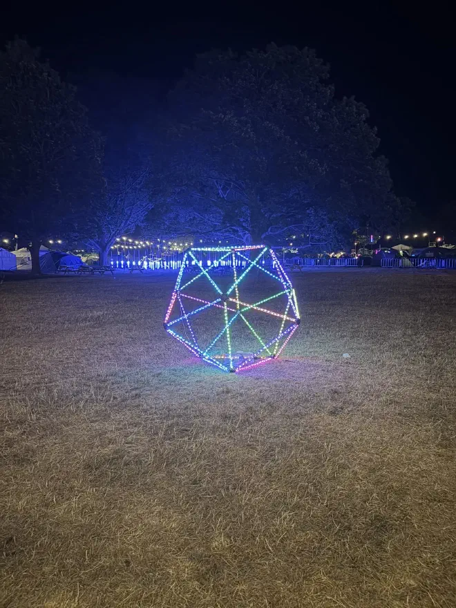 An icosahedron made of coloured LED strips sits on a grass field at night