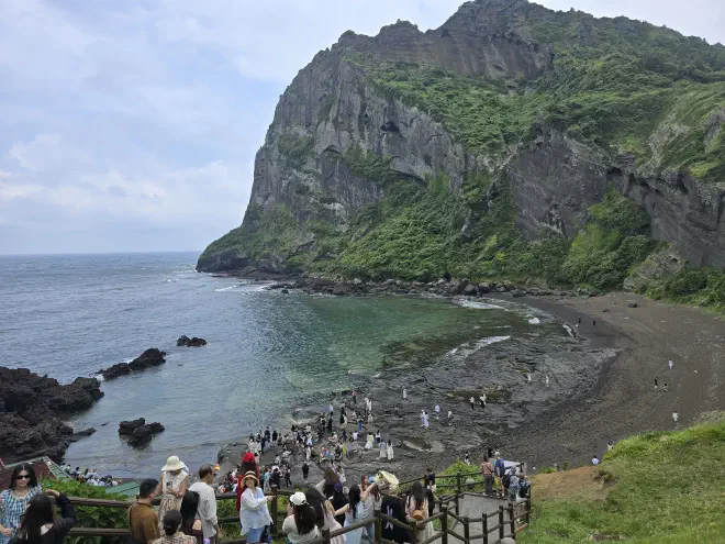 A walkway packed with tourists descends to a dark sand beach below a sheer rock face covered in fauna
