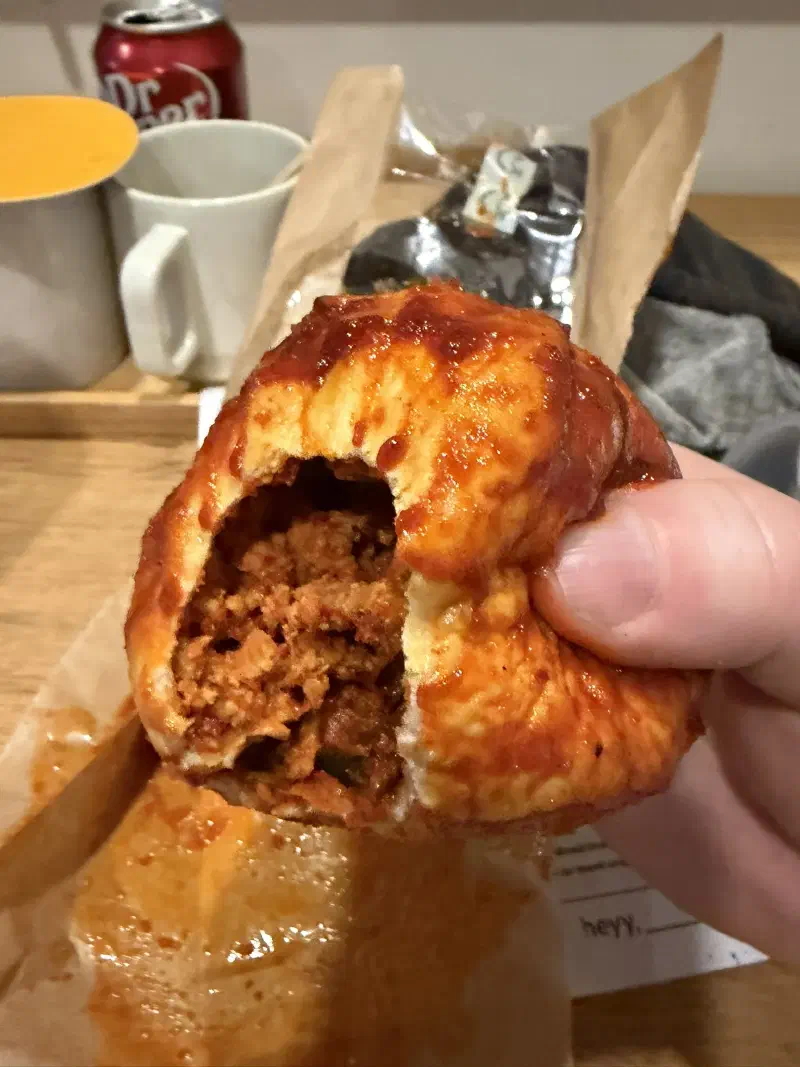 A gloved hand holds a Korean bun covered in red sauce with a bite taken from it