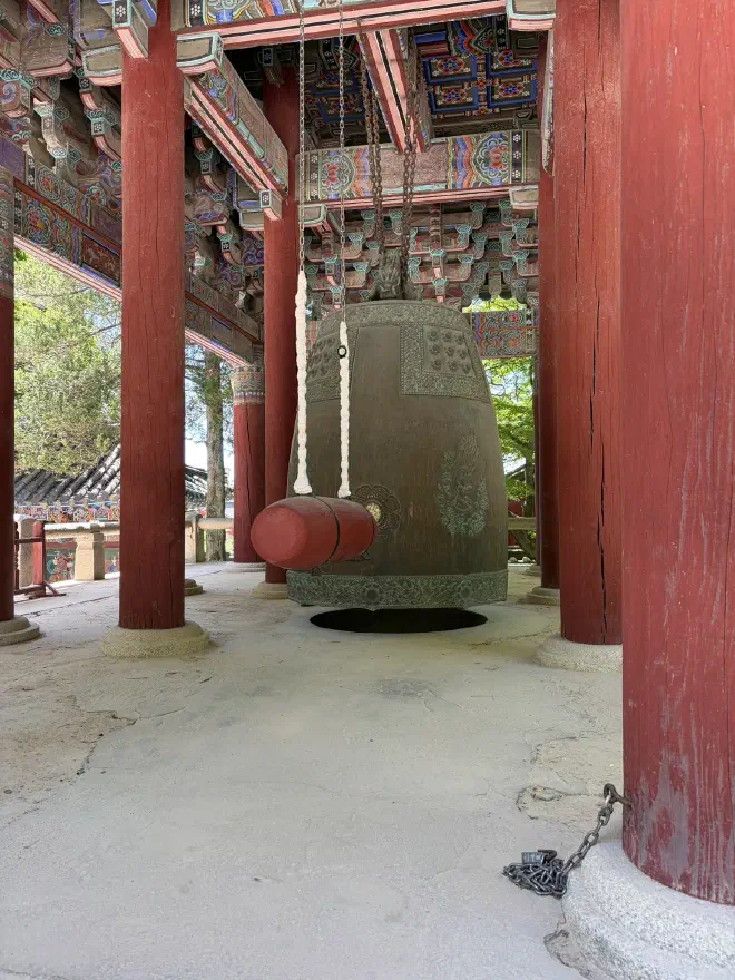 A large weathered bell hanging in a wooden building with red pillars and a painted ceiling