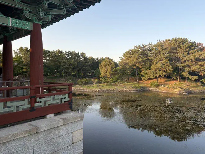 A red wooden pavillion on a stone base overlooks a stagnant pond with trees surrounding it