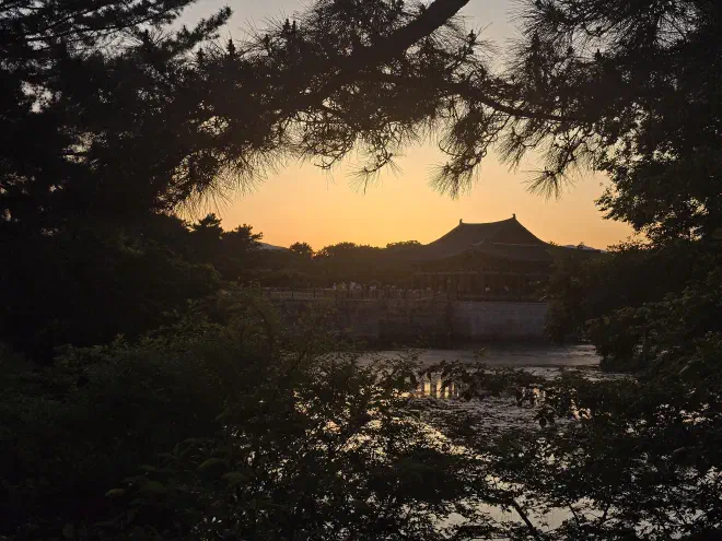 A red wooden pavillion on a stone base overlooks a pond with an orange sky at sunset