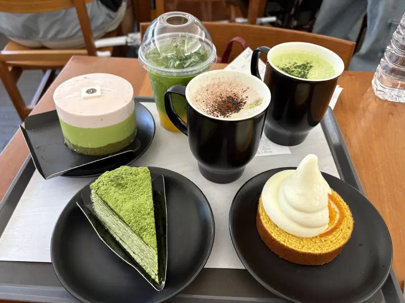 Cafe tray displaying three matcha desserts - a layered green tea cake, a mille crepe slice, and a swirled soft-serve tart—alongside two hot beverages and a cold green drink in a café setting