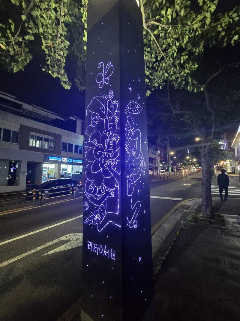 A metal post with a blue light inside it and holes drilled to display a picture of flowers and a man
