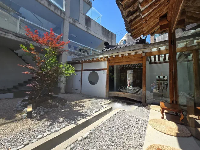 Traditional Korean courtyard blending historic hanok architecture with modern design, featuring a gravel garden with red maple tree, steam mist effect, wooden pavilion with woven floor cushions, and glass-walled contemporary building.