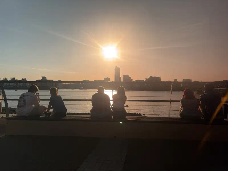 People sitting on a bench looking out over water towards a dimming sun