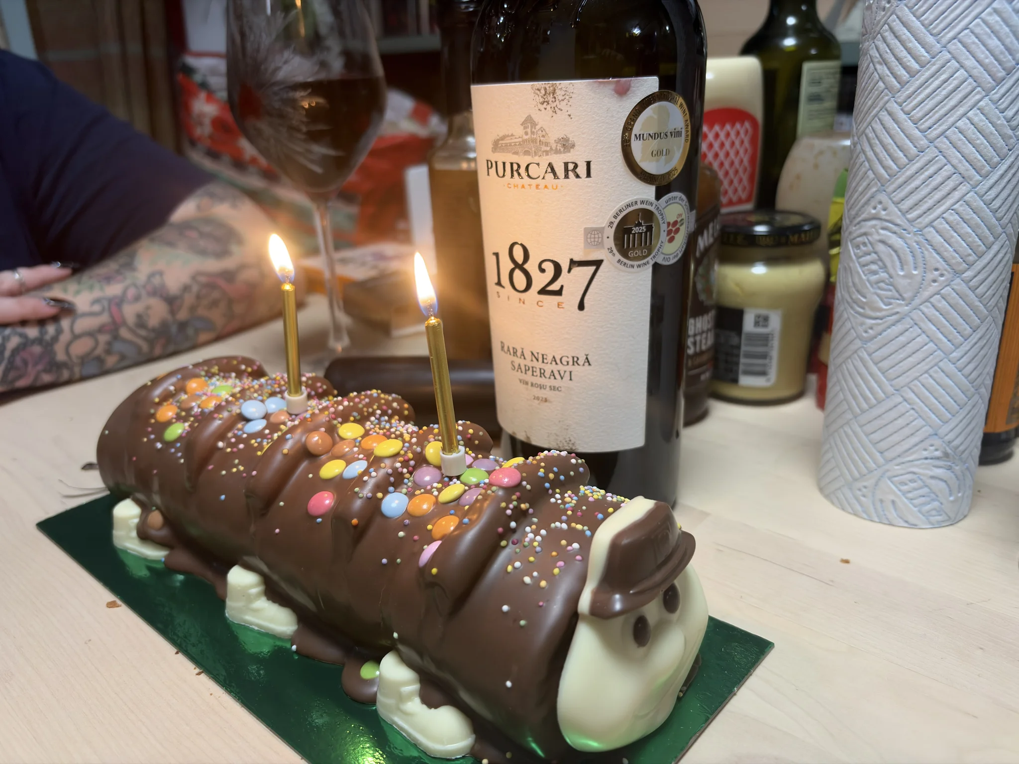 Happy Birthday to me! A caterpillar shaped cake with two lit candles on a table with a bottle of wine